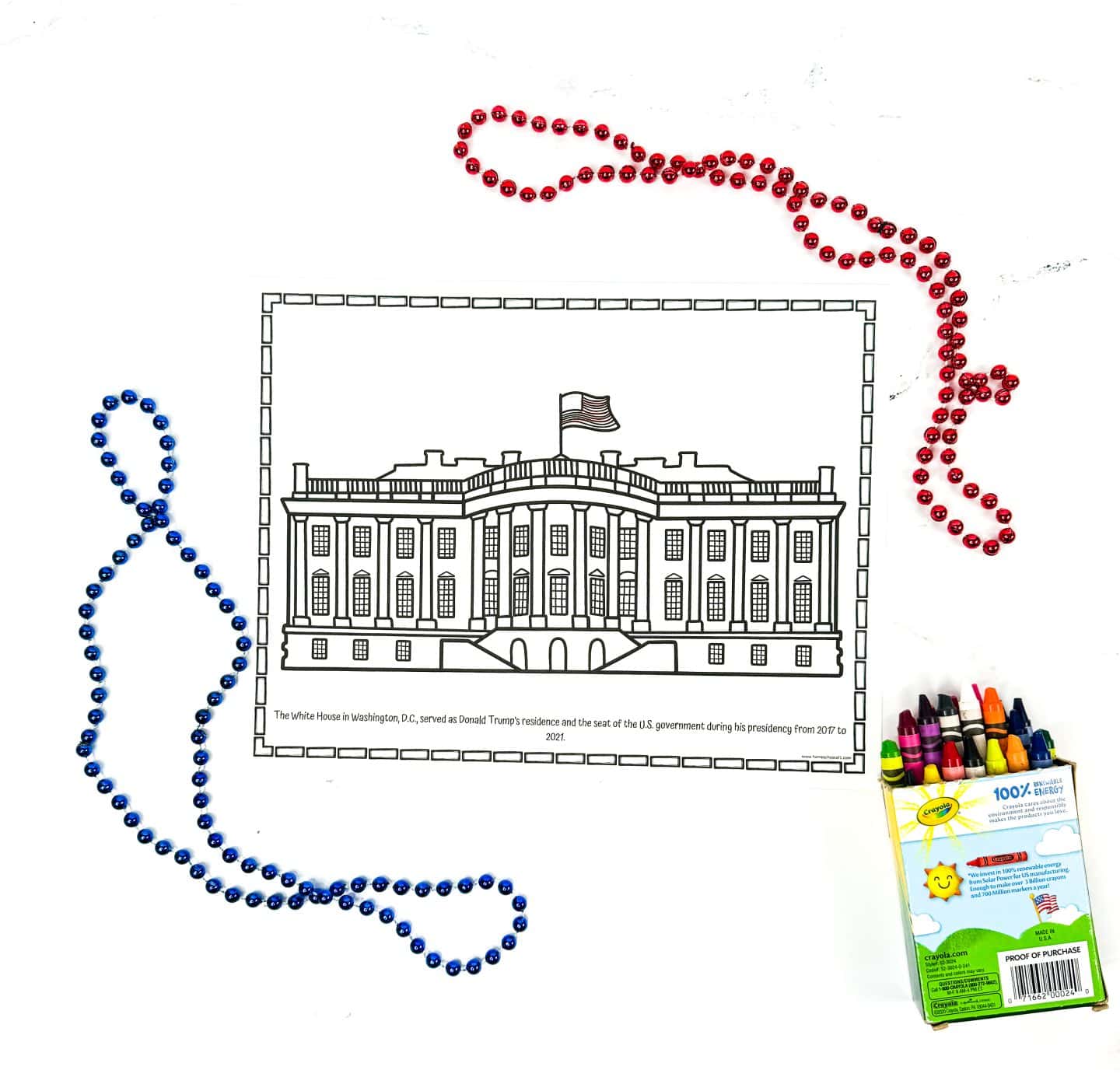A black and white coloring worksheet shows a detailed line drawing of the White House with an American flag flying on top. A caption at the bottom notes that the White House served as Donald Trump's residence and the seat of the U.S. government during his presidency from 2017 to 2021. Red and blue bead necklaces and a Crayola crayon box surround the page for a patriotic classroom activity setup.