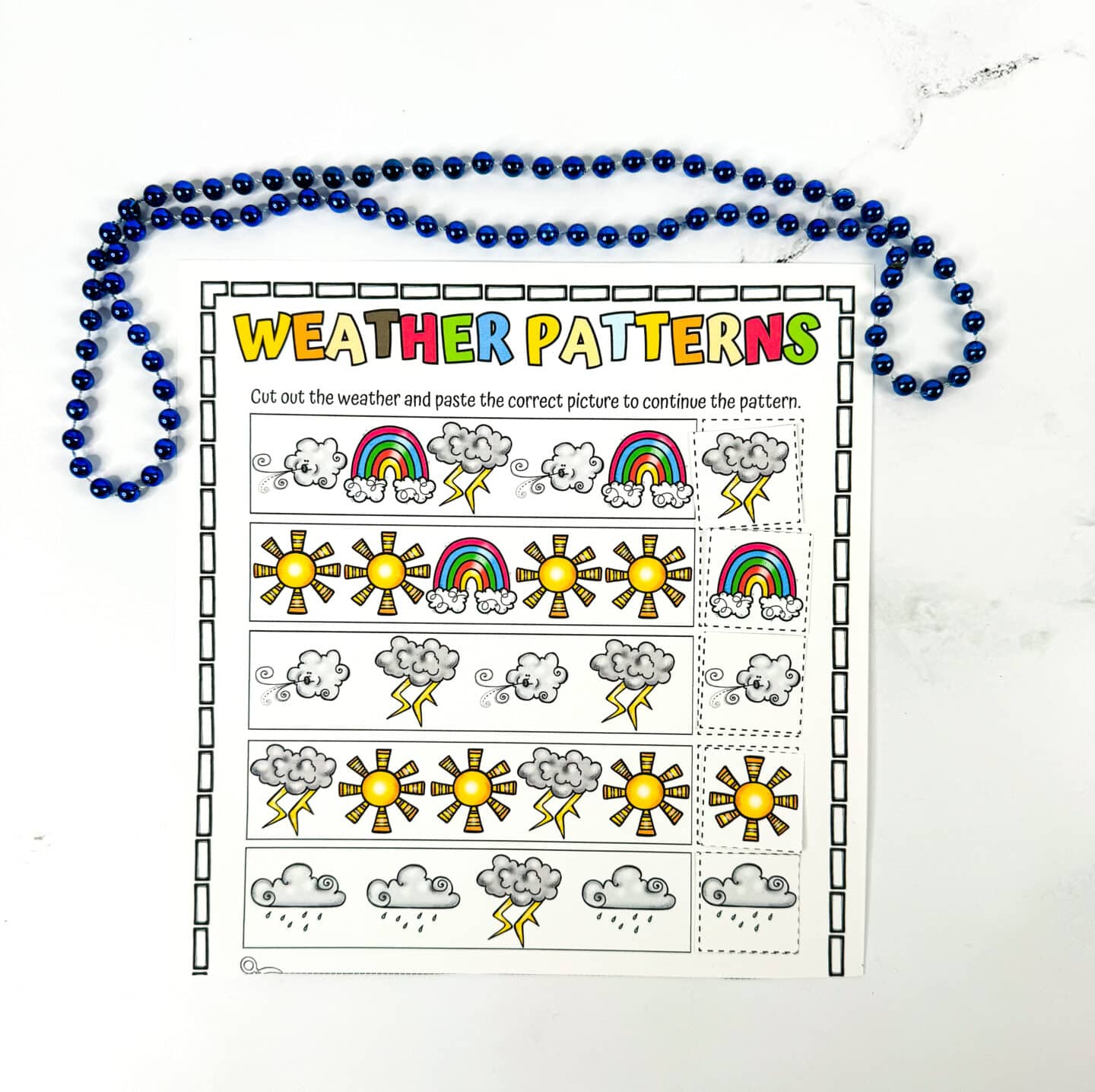 Completed weather patterns worksheet for kids showing rows of colorful weather icons—sun, rainbow, wind, clouds, lightning, and rain—with matching weather images correctly pasted to finish each pattern. The title "Weather Patterns" is in large, multicolored letters, and a blue beaded necklace is draped around the top of the sheet.
