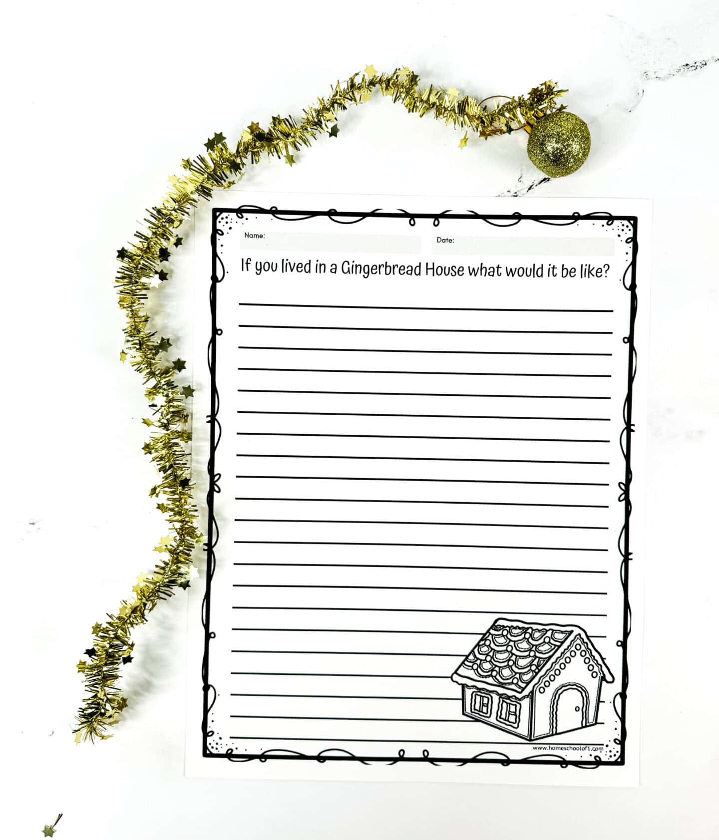 A whimsical holiday-themed writing prompt worksheet titled “If you lived in a Gingerbread House what would it be like?” features wide-ruled lines for kids to write their responses. The page is framed with a fun black border and includes a cute gingerbread house illustration in the bottom right corner. Gold star garland and a glittery ornament are placed around the page on a white background to enhance the festive vibe.

