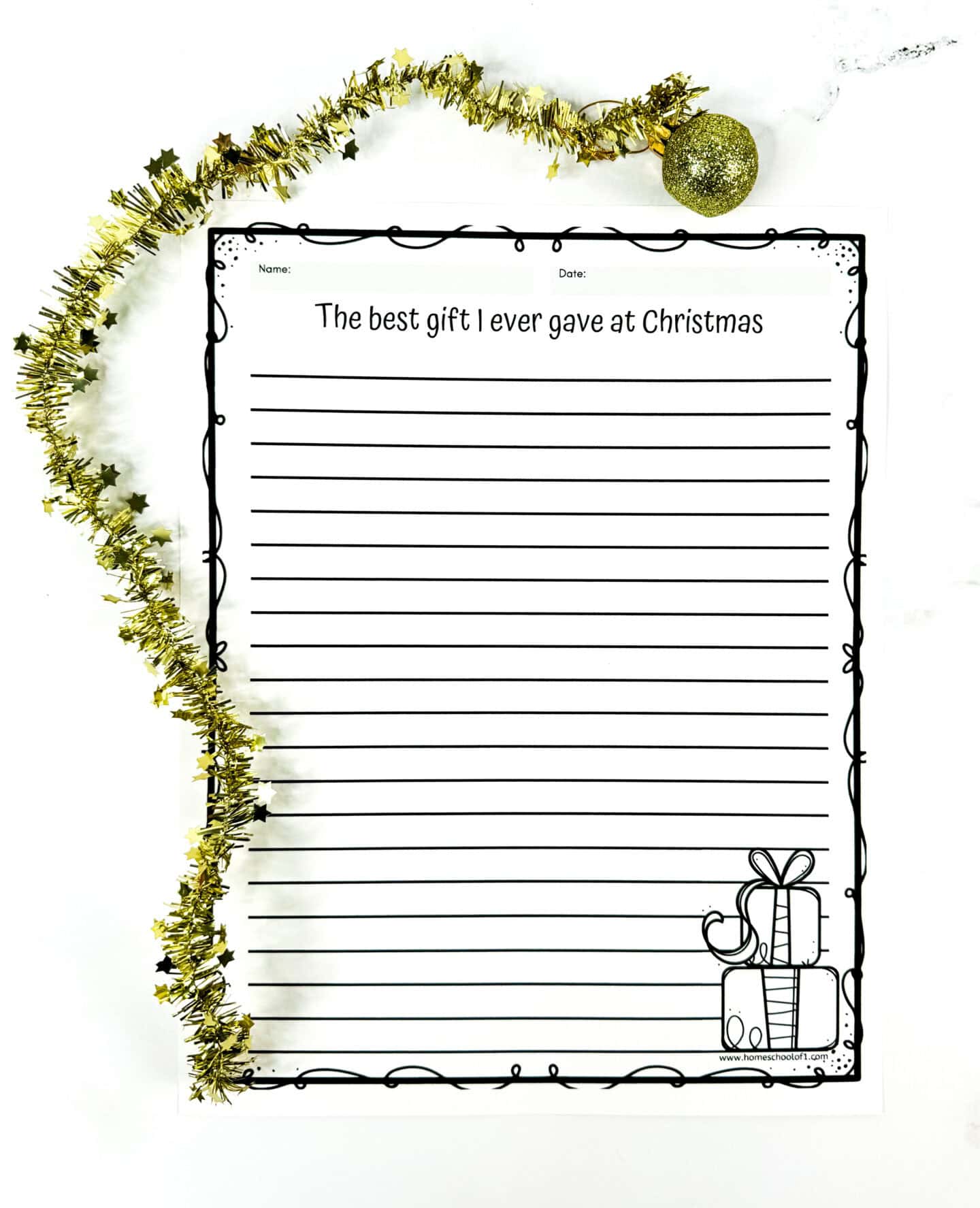 A holiday-themed writing prompt worksheet titled “The best gift I ever gave at Christmas” features lined space for children to write their response. The page has a decorative black border and a simple illustration of stacked presents tied with bows in the bottom right corner. Gold star garland and a glittery ornament frame the top of the page, adding festive cheer.

