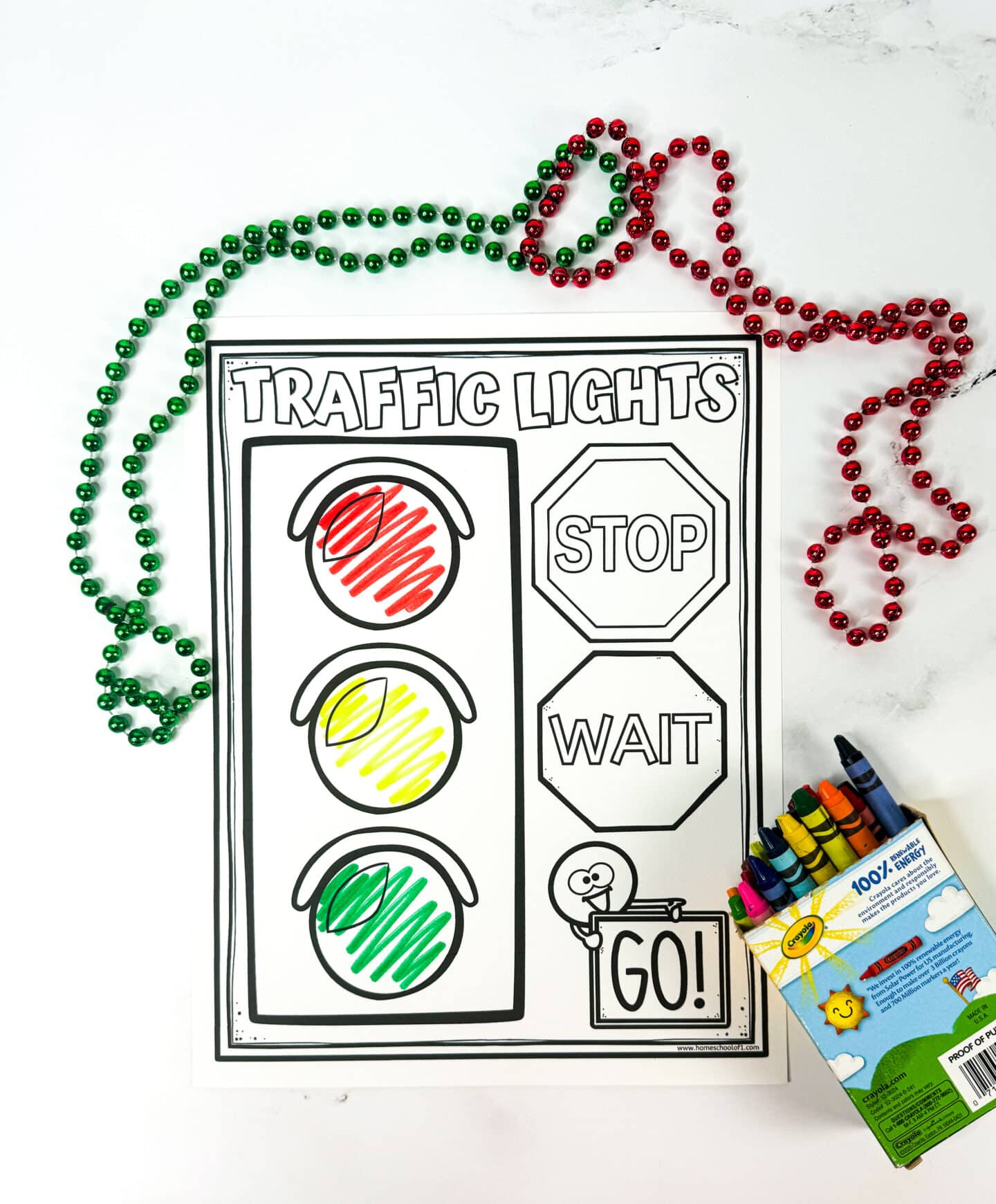 This image shows a printable traffic light activity sheet for kids, partially colored with red, yellow, and green crayon scribbles in the respective traffic light circles. On the right side, labeled shapes include a red stop sign with "STOP," a yellow hexagon with "WAIT," and a green rectangle with "GO!" held by a stick-figure character. Red and green Mardi Gras-style beads are draped around the page, and a Crayola crayon box sits nearby.