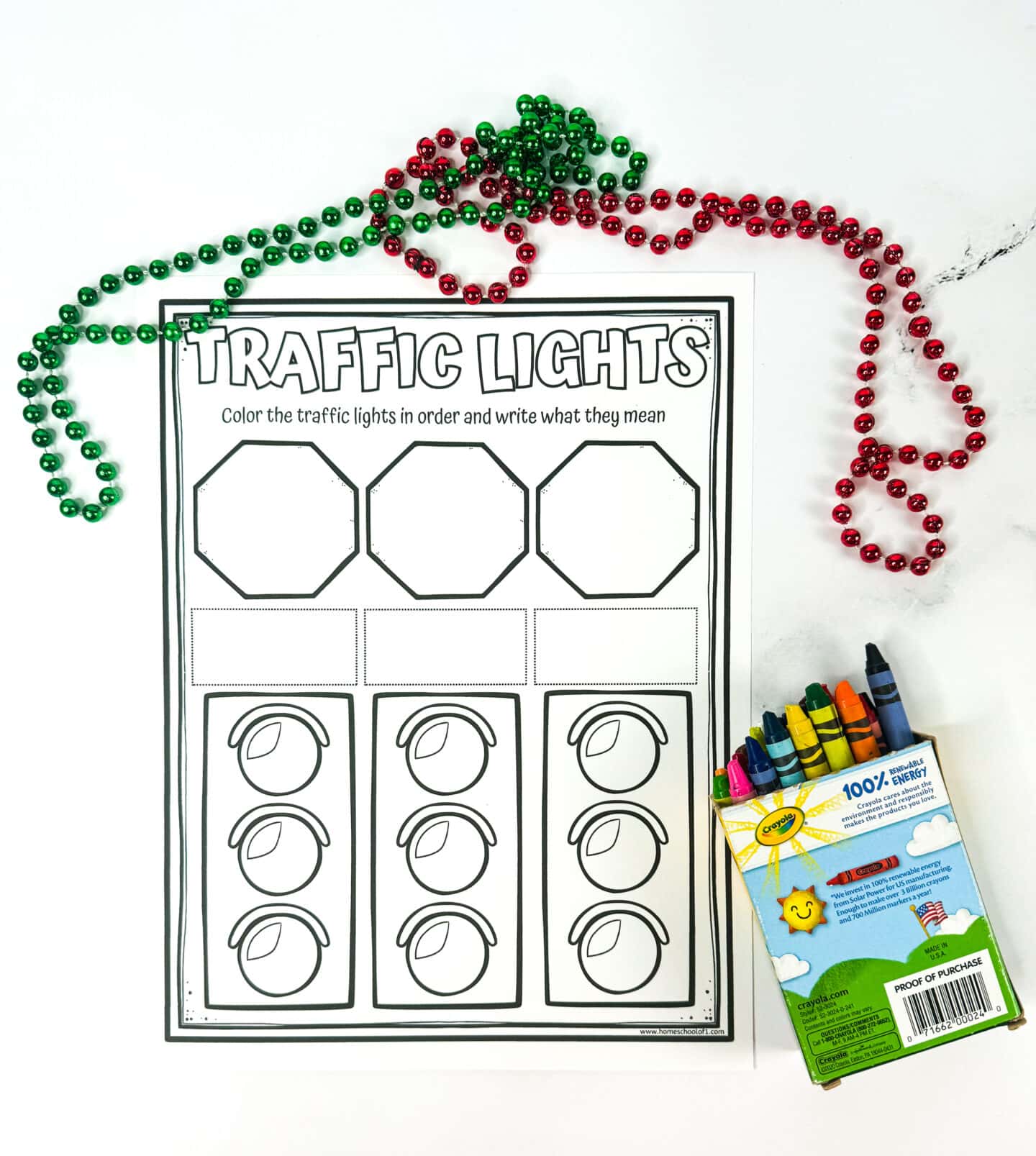 A blank traffic light-themed worksheet for kids, with three octagons at the top labeled "Color the traffic lights in order and write what they mean." Below are three sets of uncolored traffic lights and empty boxes for writing. Green and red bead necklaces are arranged decoratively around the sheet, and a colorful box of Crayola crayons sits nearby.