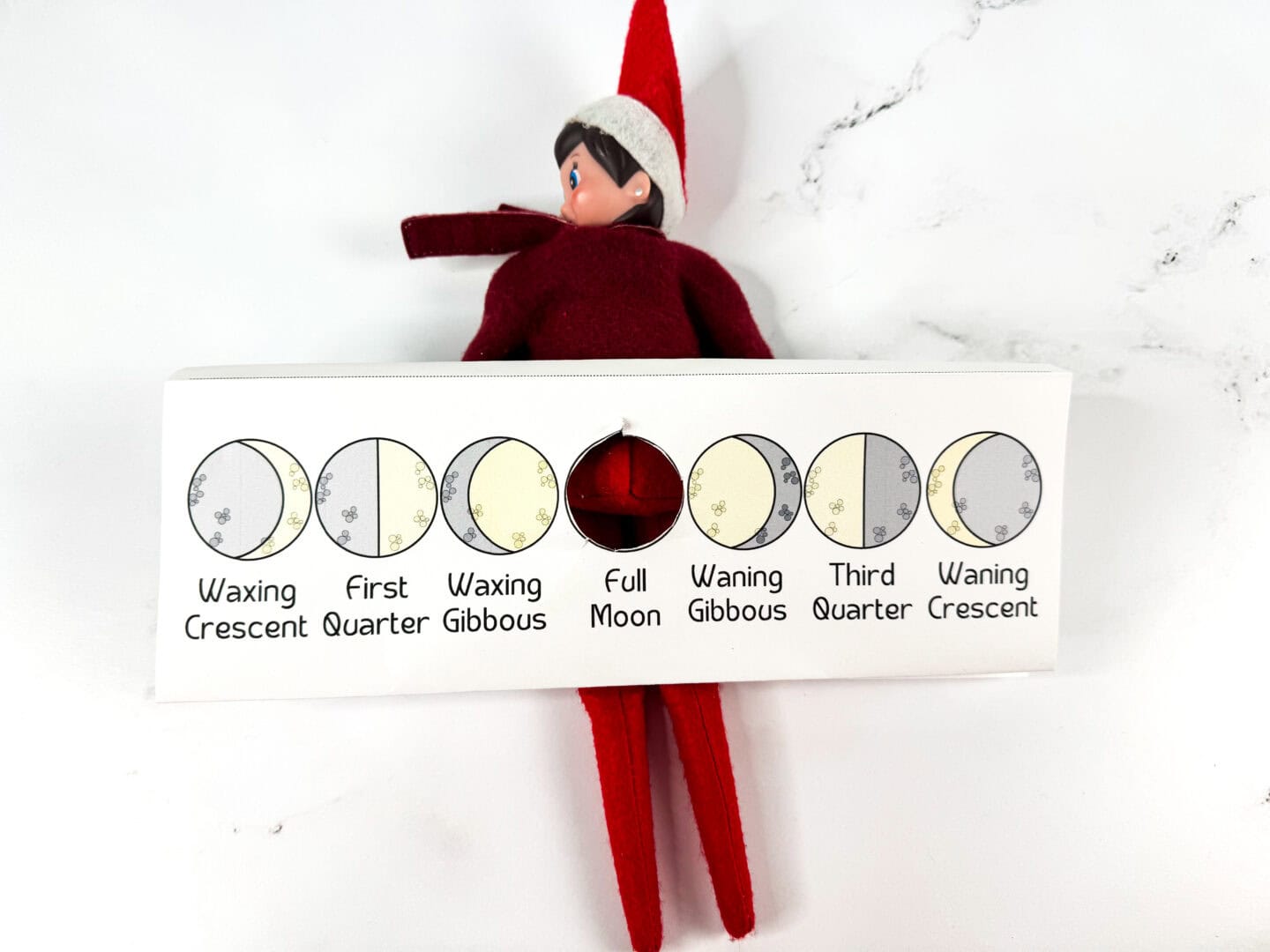 An Elf on the Shelf lies behind a folded moon phases printable with a circular cutout over the “Full Moon” phase, revealing part of the elf’s red outfit. The chart includes labeled illustrations of all seven phases: Waxing Crescent, First Quarter, Waxing Gibbous, Full Moon, Waning Gibbous, Third Quarter, and Waning Crescent. The elf adds a playful twist to this educational science craft.

