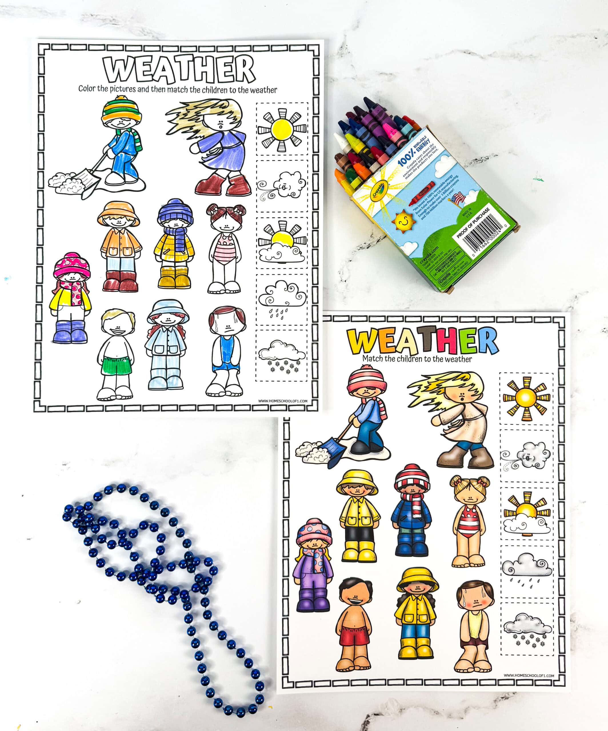 Two printable weather-themed worksheets for kids show children dressed in various outfits suitable for different weather types, like snow, rain, sun, and wind. One worksheet is partially colored, while the other is fully colored, and both include matching weather icons like suns, clouds, and snowflakes. A box of crayons and a blue beaded necklace rest on the white marble surface alongside the pages, emphasizing a fun and creative learning activity.