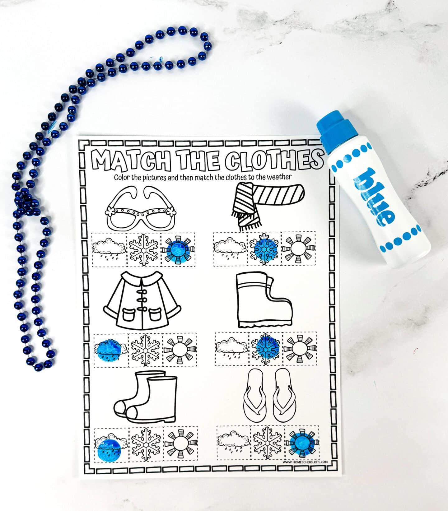 A black-and-white children’s worksheet titled "Match the Clothes" features illustrations of weather-appropriate clothing like a scarf, sunglasses, raincoat, boots, and flip-flops. Kids are instructed to color the items and match them to weather symbols; some matches are already made with blue bingo marker dots placed on snowflake and sun icons. The scene also includes a blue beaded necklace and a blue dot marker labeled “blue,” all on a white marble background.