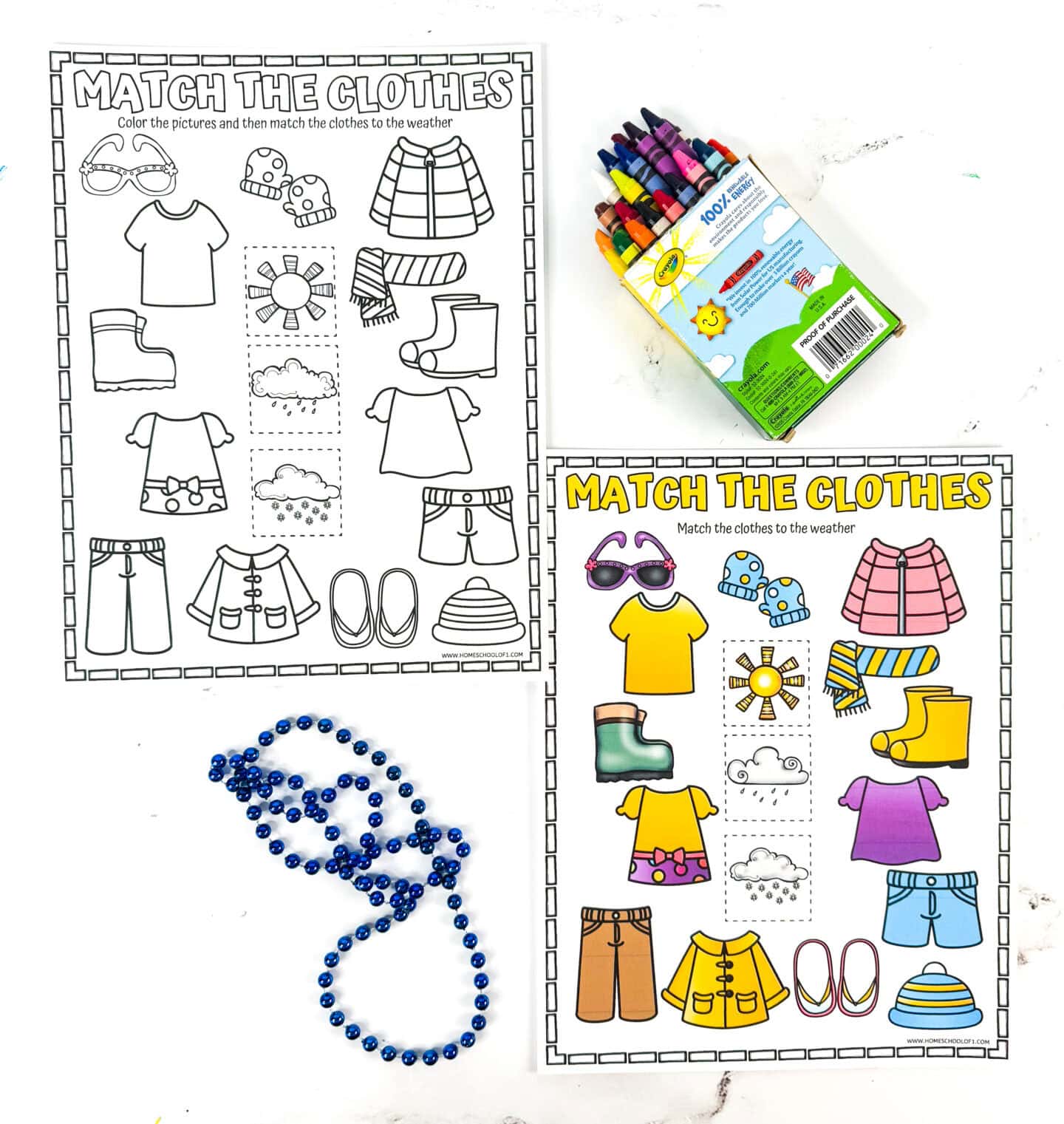 Two printable worksheets titled "Match the Clothes" feature a variety of clothing items such as shorts, rain boots, a jacket, flip-flops, and a winter coat, with corresponding weather icons including sun, clouds, and snow. One worksheet is in black and white for coloring, while the other is fully colored and completed. A box of crayons and a blue beaded necklace are placed beside the worksheets on a white marble background, highlighting a fun and educational weather-themed activity.