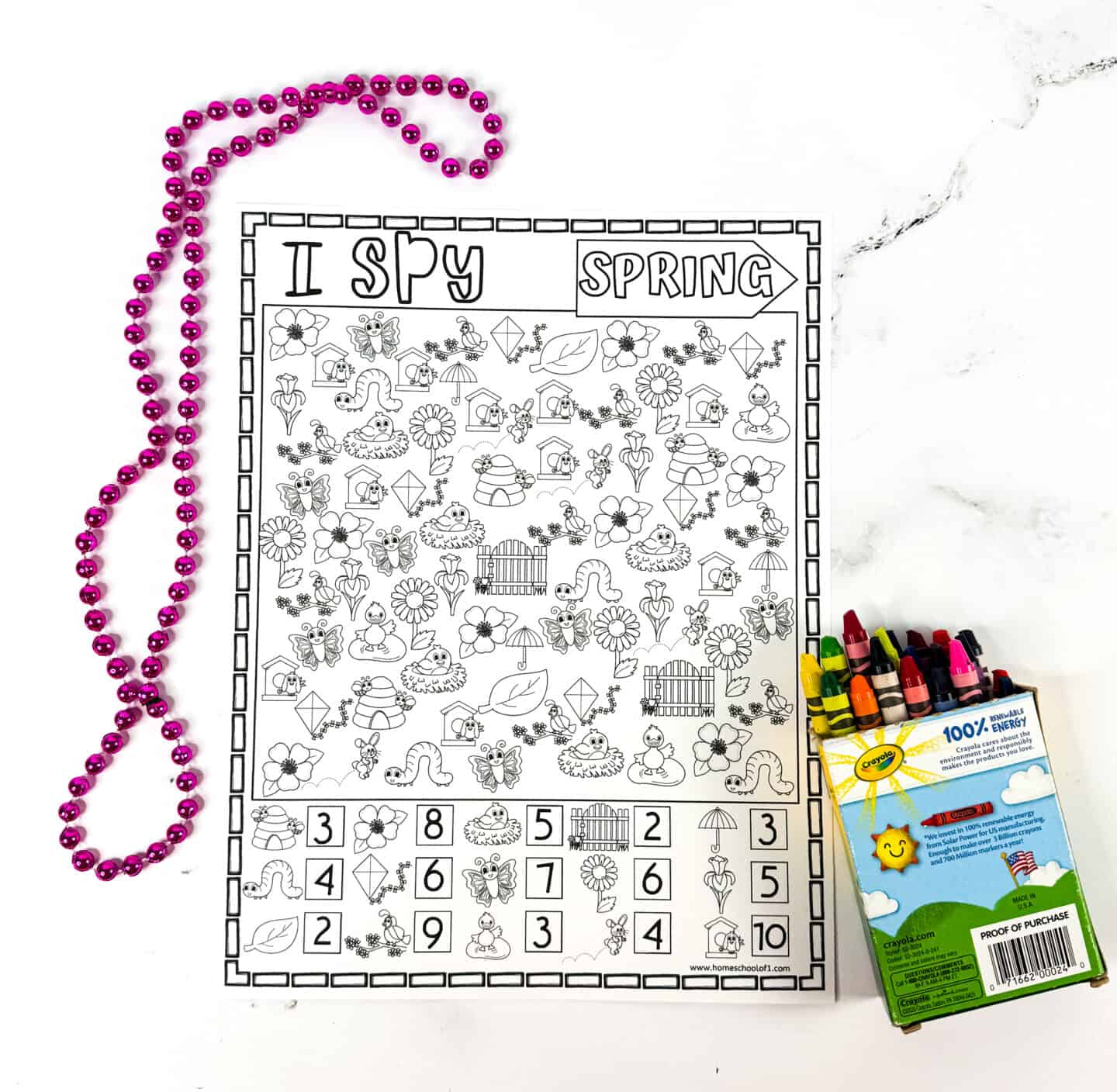 Black and white printable "I Spy Spring" worksheet featuring spring-themed icons like flowers, birds, kites, butterflies, rain boots, birdhouses, and garden fences, with a counting key at the bottom. A pink beaded necklace is looped to the left, and a box of Crayola crayons sits to the right on a white marble surface.

