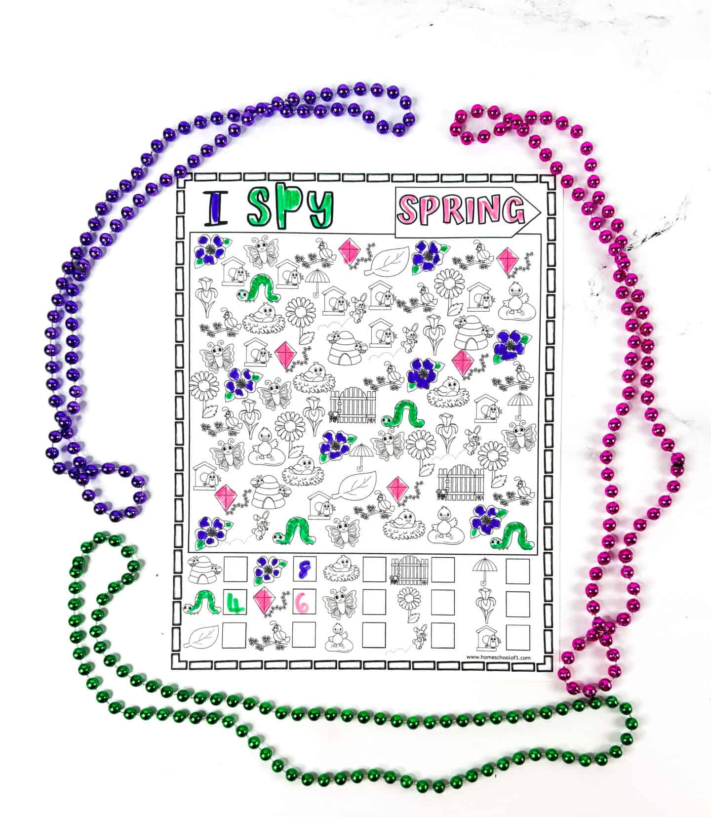 Spring-themed "I Spy" printable partially colored in with green, pink, and purple markers, featuring items like flowers, butterflies, kites, and worms. It’s surrounded by colorful beaded necklaces in purple, pink, and green, laid out on a white marble background.