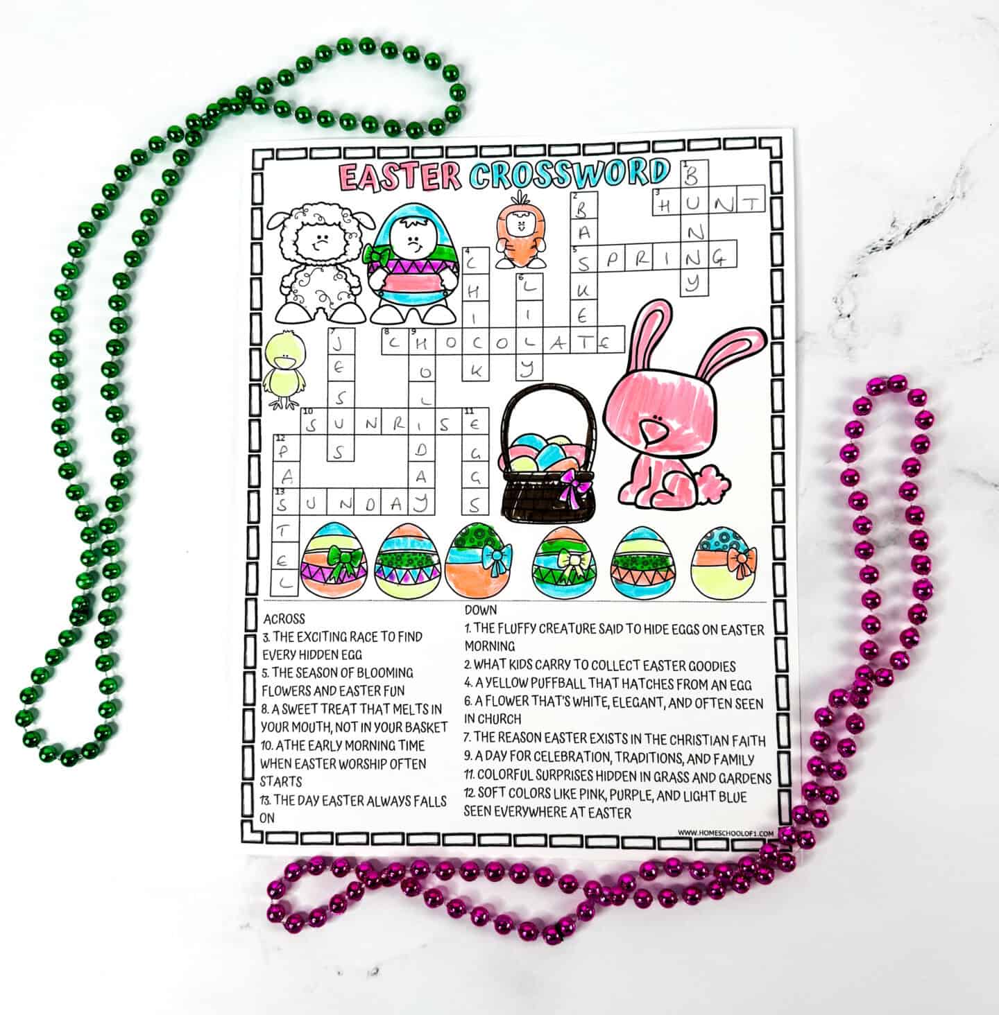 Colorful Easter crossword puzzle page partially filled in, with words like "bunny," "chocolate," "sunrise," and "Sunday." The page features cheerful illustrations including a pink bunny, lamb, Easter basket, and brightly decorated eggs. Surrounded by green and pink beaded necklaces arranged playfully on a white marble background.