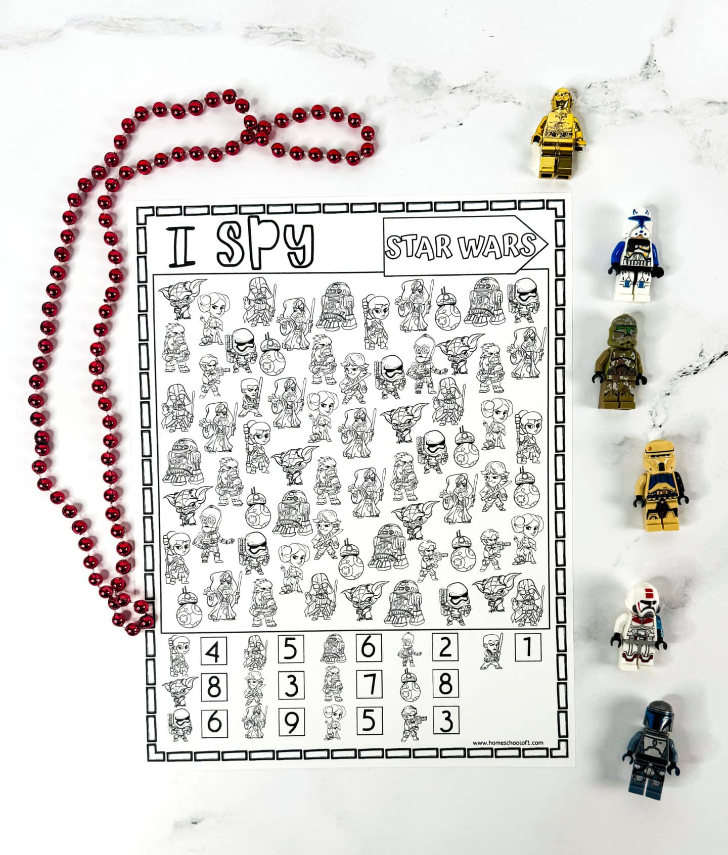 A black and white “I Spy Star Wars” activity sheet featuring various cartoon Star Wars characters and a key at the bottom showing how many times each character appears. The page is surrounded by LEGO Star Wars minifigures, including a golden C-3PO, clone troopers, and others, along with a red beaded necklace shaped into a bow on a white marble background.