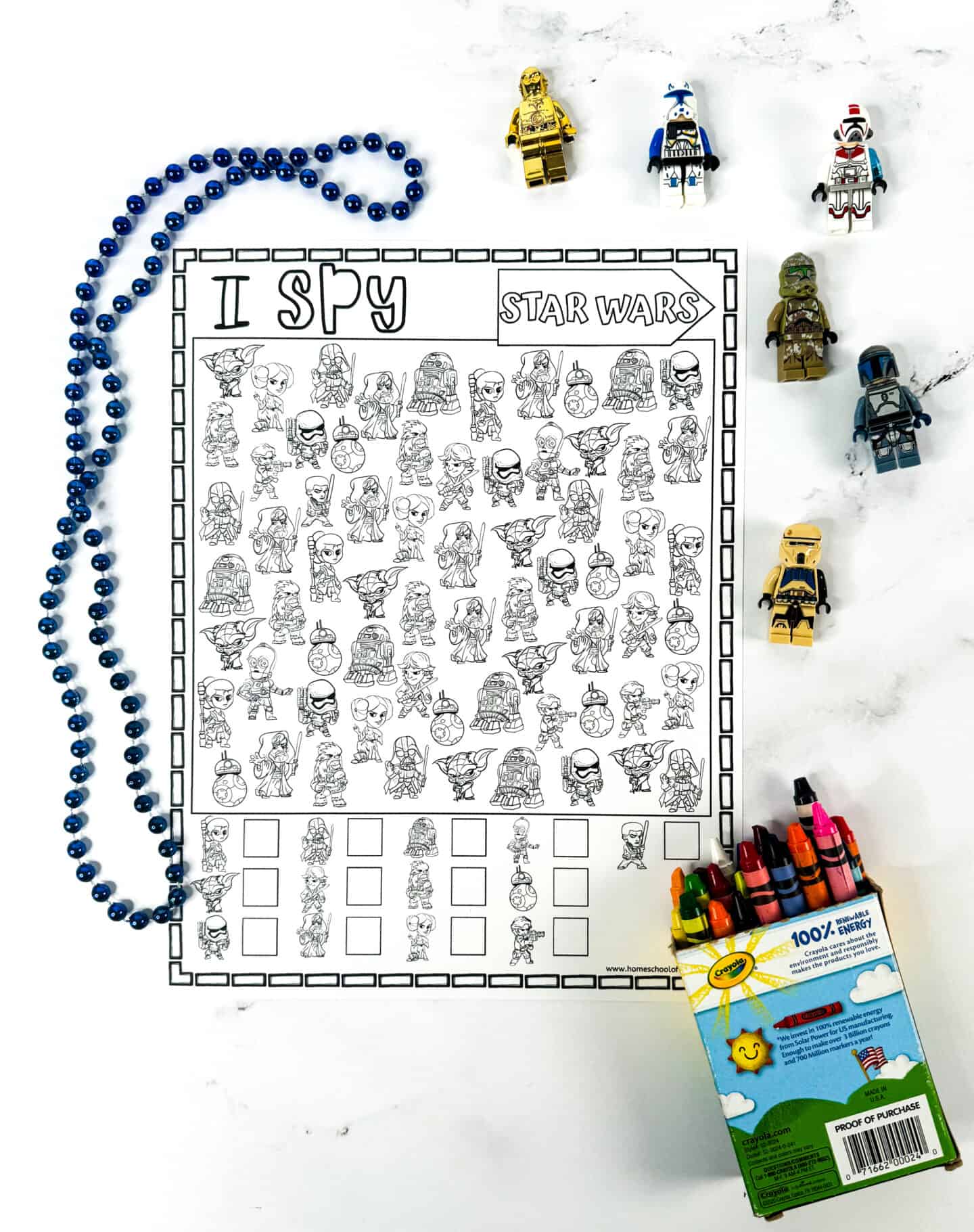 A black and white "I Spy Star Wars" activity sheet featuring cartoon-style characters from the Star Wars franchise. Surrounding the sheet are various LEGO Star Wars minifigures, a string of blue beads, and an open box of Crayola crayons, suggesting a fun and creative activity for kids.