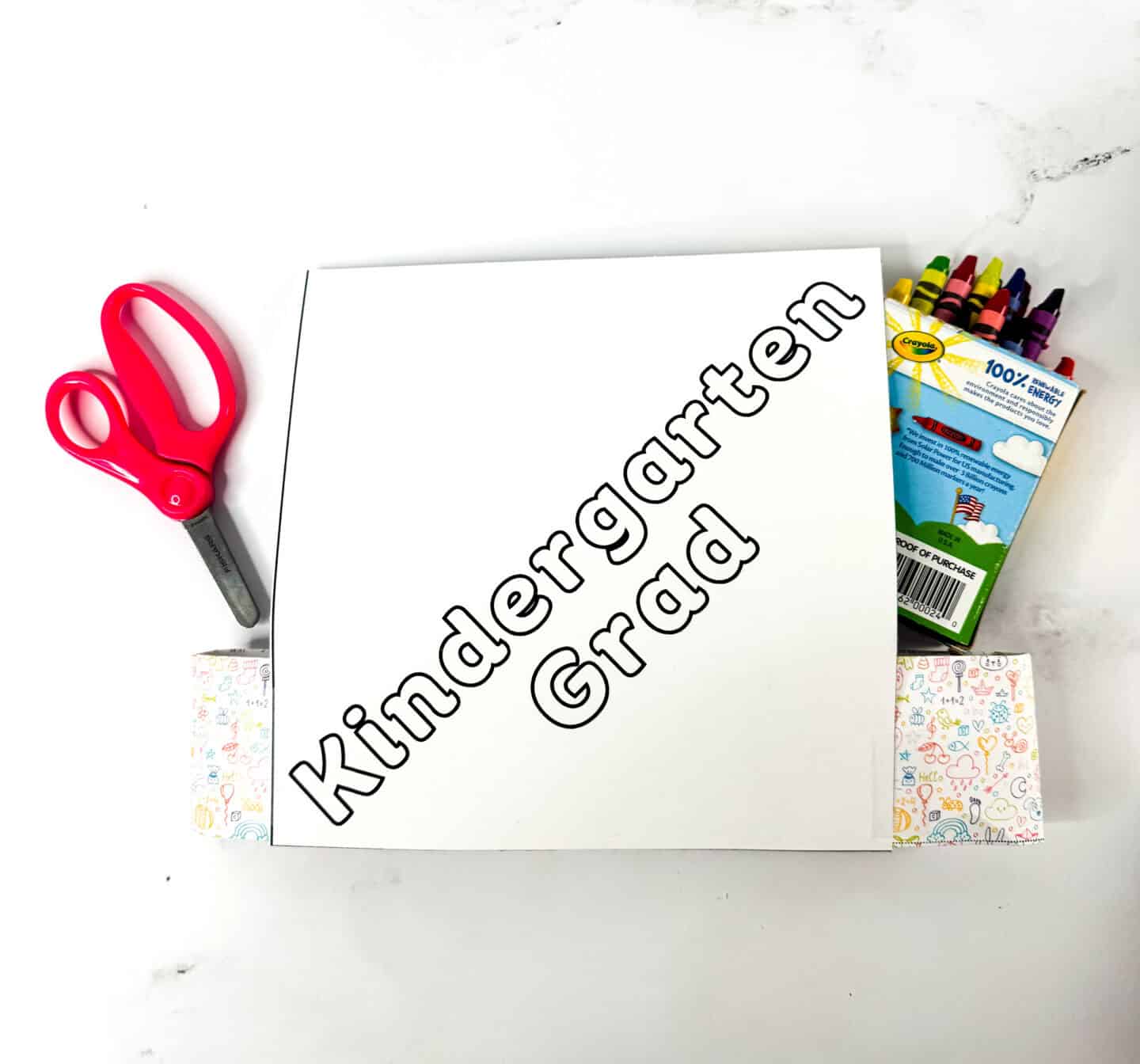 A square paper with the words "Kindergarten Grad" in large outlined letters, ready to be colored, with a pair of child-safe red scissors on the left and a box of Crayola crayons on the right.