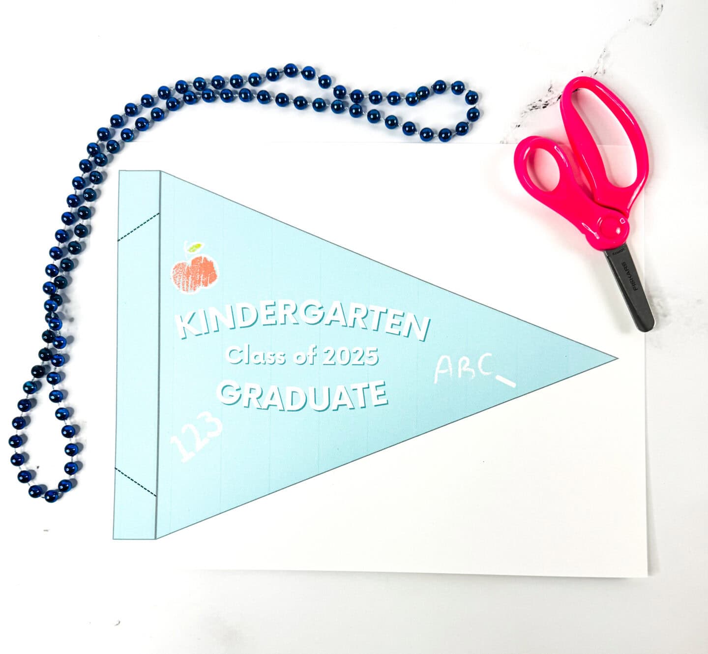 A printable light blue pennant labeled "Kindergarten Class of 2025 Graduate" with chalk-style drawings of "123", "ABC", and an apple, placed between a pair of safety scissors and a navy blue beaded necklace.