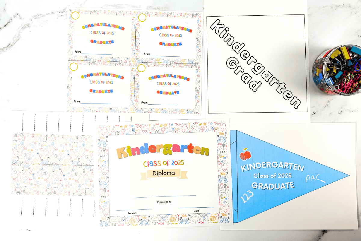 Flat lay of printable kindergarten graduation items including four congratulatory cards, a large diploma, a blue pennant that reads "Kindergarten Class of 2025 Graduate", and a "Kindergarten Grad" sign next to a jar filled with paperclips and binder clips.