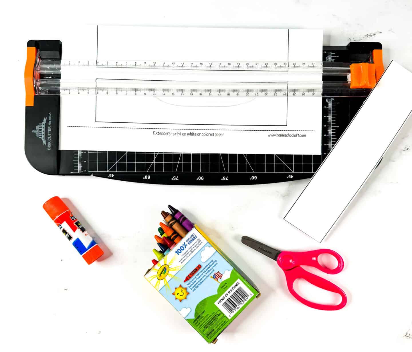 Halloween crown extender strips being trimmed with a paper cutter, shown alongside glue stick, pink scissors, and a box of crayons.