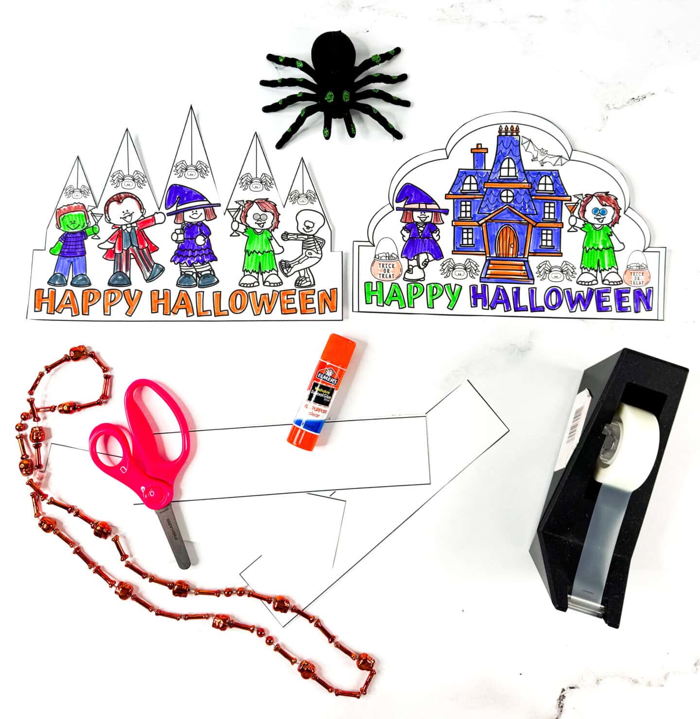 Flat lay of a Halloween crown craft setup including cut-out crown designs, scissors, glue stick, tape, toy spider, and orange beaded necklace.