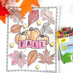 Printable fall leaf coloring page with pumpkins, colored leaves, and the word “FALL” in block letters. Surrounded by crayons and fake fall leaves.
