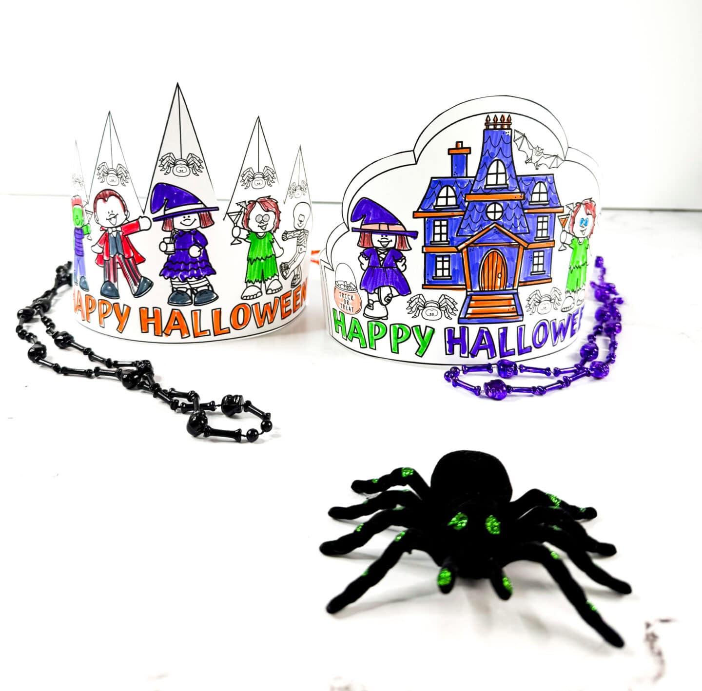 Two Halloween crown printables fully colored and assembled, featuring trick-or-treat characters and a haunted house, with Halloween necklaces and a toy spider in front.