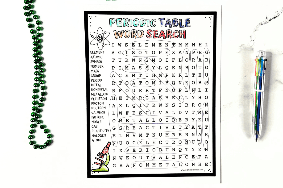 Periodic Table Word Search worksheet on a desk with pen and green beads, featuring chemistry vocabulary like mass, group, and reactivity.