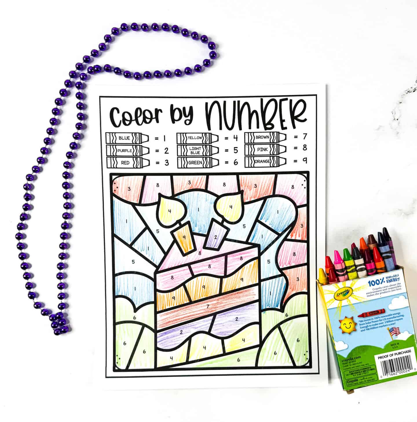 Birthday color by number sheet featuring a layered birthday cake with candles, colored with crayons and placed beside purple beads.