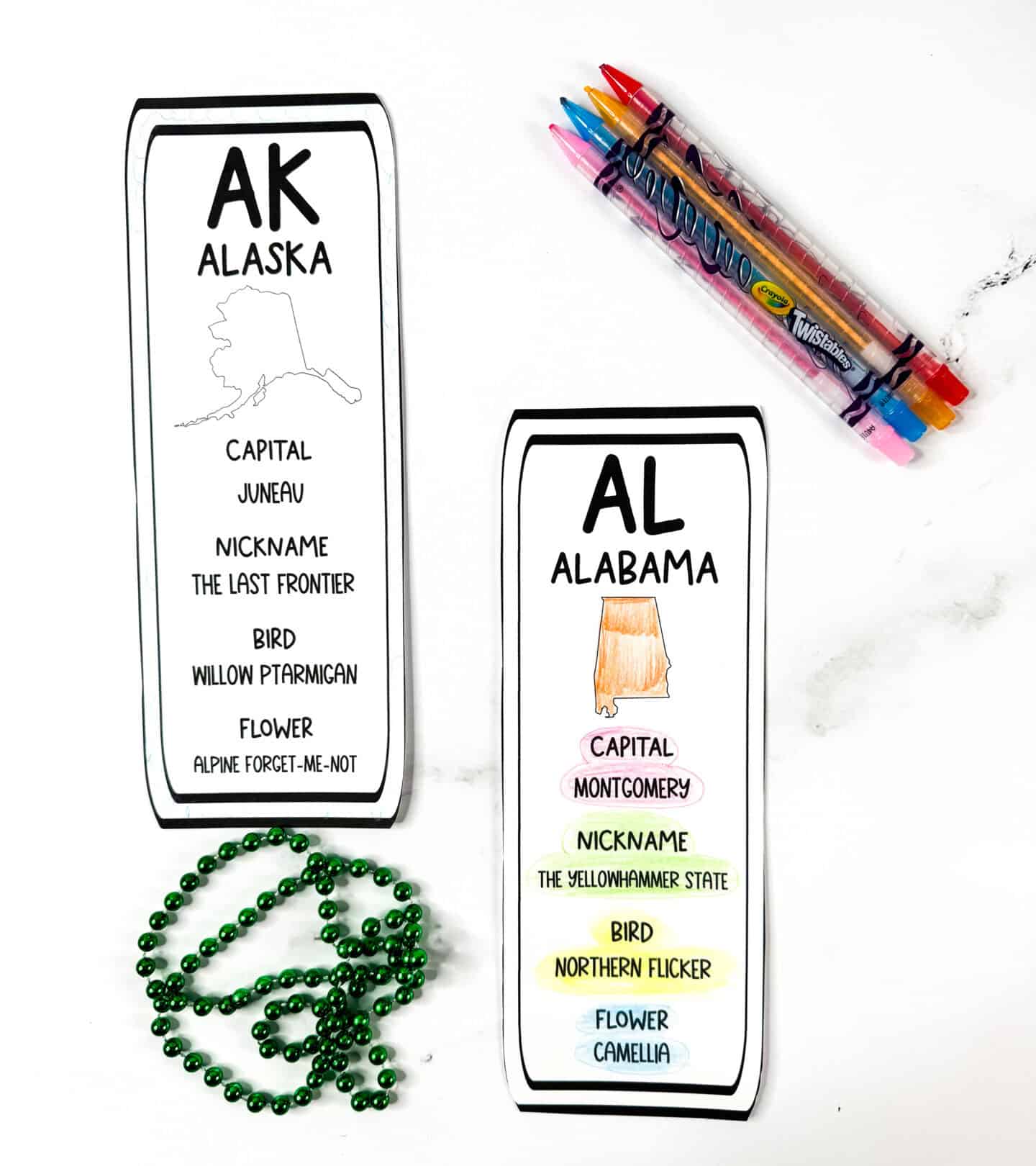 Black and white Alaska bookmark next to a colored Alabama state bookmark featuring an orange state shape and pastel highlights on key facts. Crayola Twistable crayons and green bead garland used for styling.