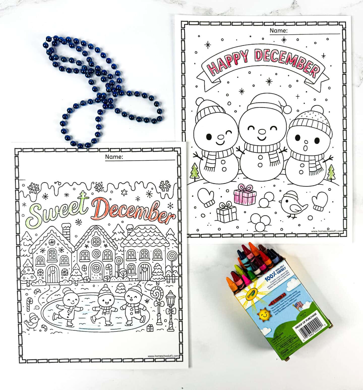 Two December coloring sheets, one showing happy snowmen under “Happy December” and one gingerbread village labeled “Sweet December”—arranged with crayons and blue beads.
